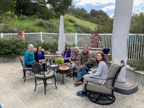 Several individuals sit outside on a patio in outdoor furniture with green vegetation in the background.